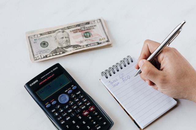 Hand writing on a notepad next to a black calculator and a small stack of U.S. dollars